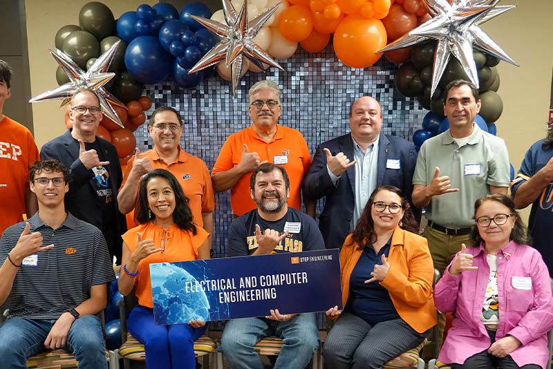 Group of attendees pose together during a UTEP Engineering homecoming event while holding a large “Electrical and Computer Engineering” banner, seated and standing in rows in front of a decorative balloon backdrop and making hand gesture signs during the departmental group photo.